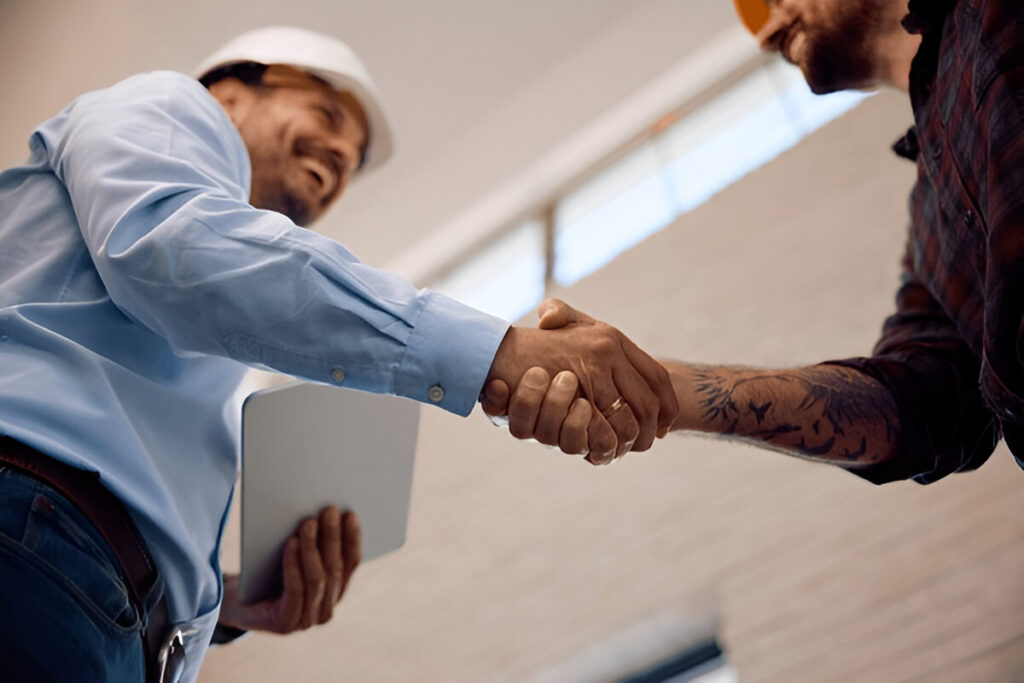 Close-up of a construction site engineer in a white hard hat shaking hands with a business partner while holding a tablet.