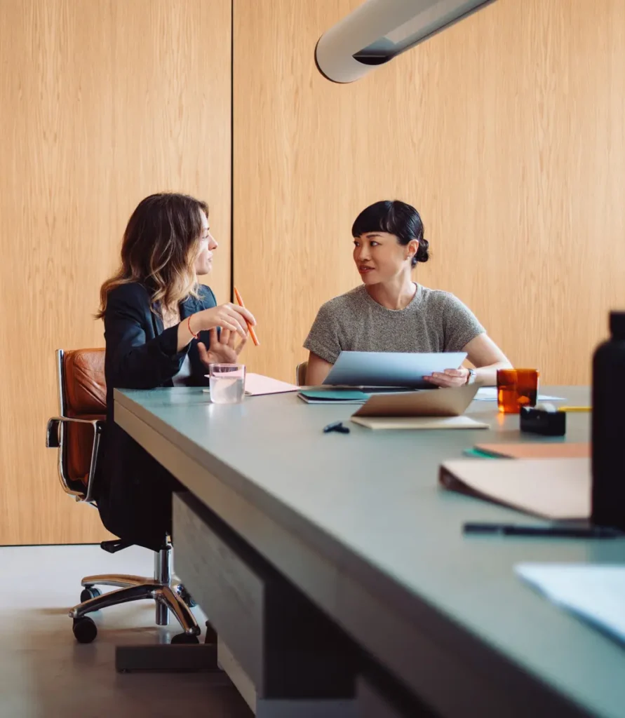 Two diverse corporate professional women sitting at a conference table reviewing business documents and planning a strategy.